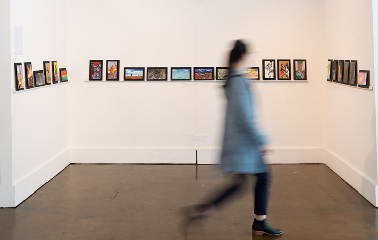 A blurred figure walks in front of a wall of 9 by 5 artworks.