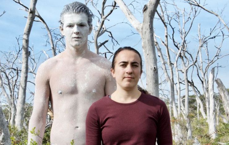 Two performers, one covered in white body paint, in front of bare trees