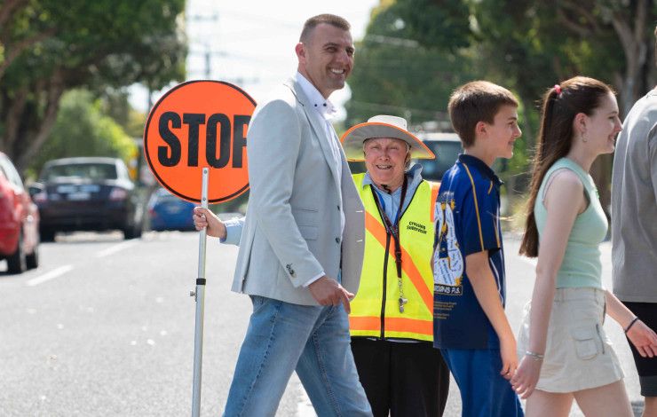 People crossing a pedestrian crossing in front of a supervisor with an orange stop sign.