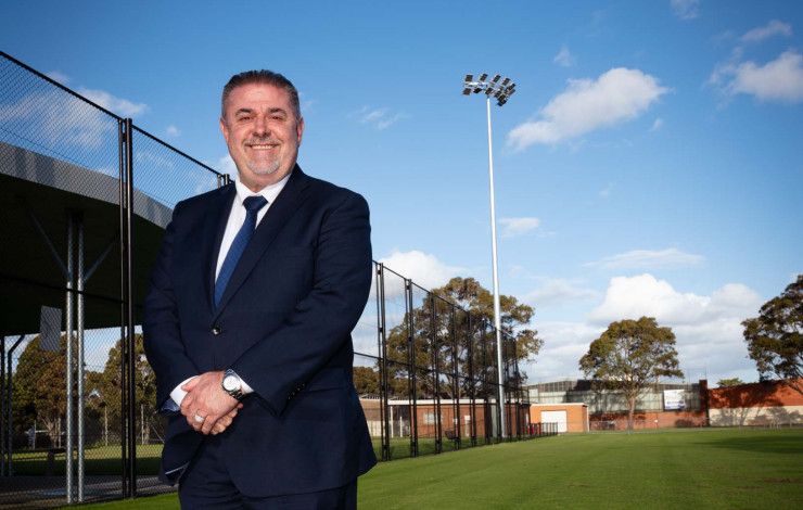 Man in a dark suit stands on a sports ground under a large light tower