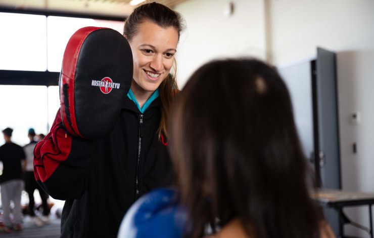 A smiling woman holds a boxing pad in front of someone standing with their back to the camera