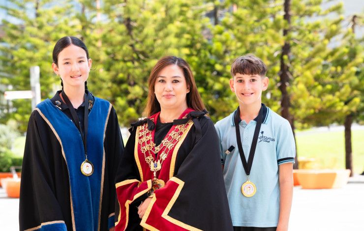 Two smiling children stand either side of a woman dressed in red and black robes