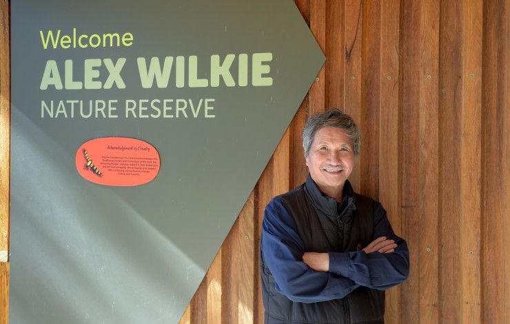 A smiling man with his arms crossed next to a sign that reads Welcome Alex Wilkie Nature Reserve