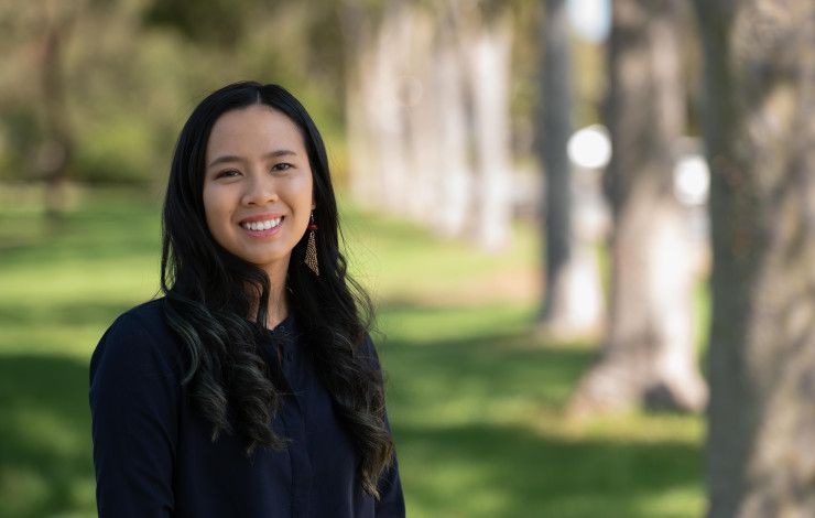 A woman with long dark hair smiles at the camera while standing in a park location
