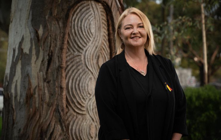 A smiling woman dressed in black stands in front of an Aboriginal message tree.