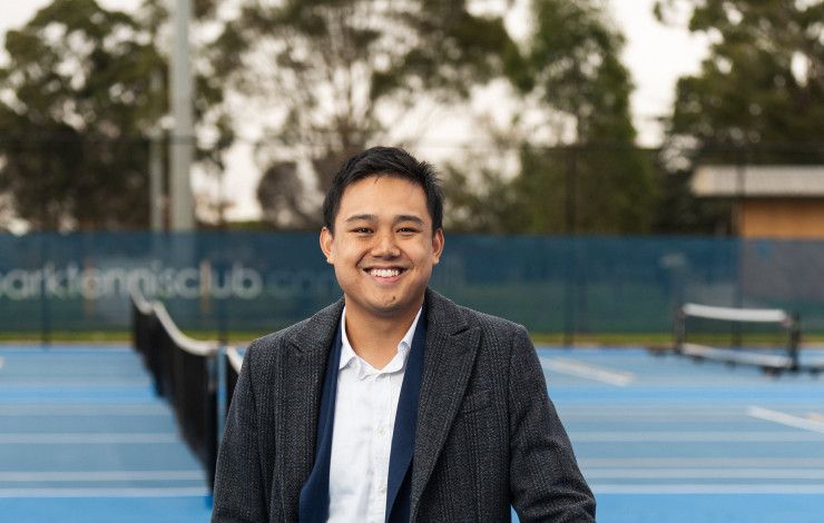 A smiling man leans on a tennis net.
