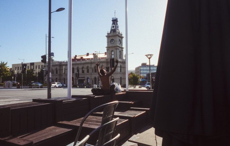A man sits in the sunshine with his hands raised to the air across the street from a historical building and clock tower
