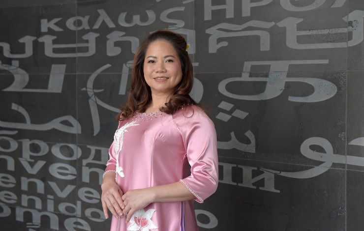 A smiling woman dressed in a pink silk dress in front of a black wall covered in grey lettering