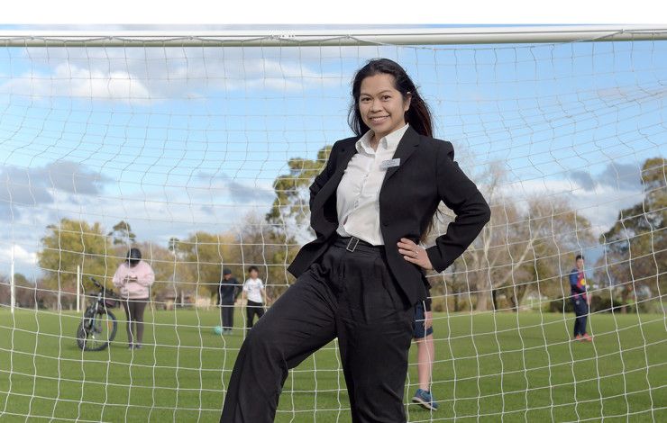 A smiling woman dressed in a black suit and white shirt stands in front of a soccer goal with one hand on her hip