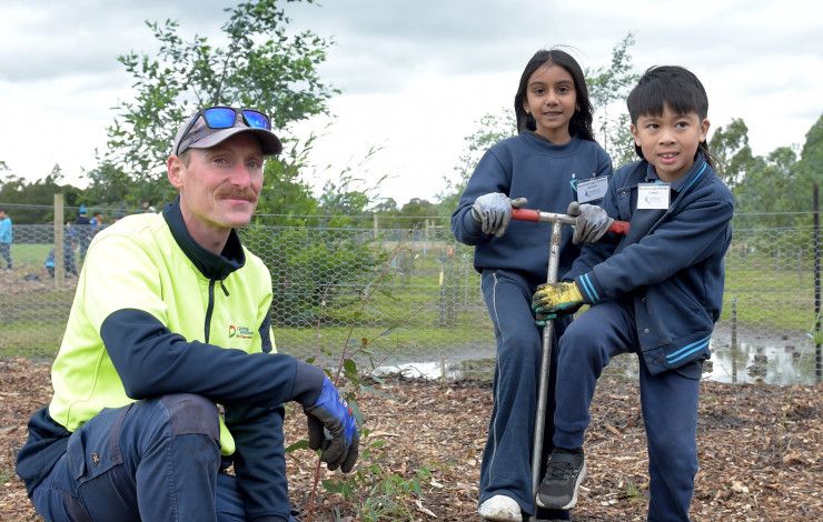 A man crouches next to two children who are helping him plant a tree