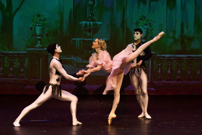A female ballet dancer in pink costume dances with two male dancers dressed as harlequins