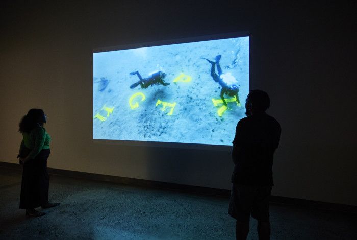 People watching a projection of people scuba diving near giant yellow letters.