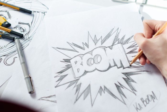 A person sketching with a pencil. The words Boom and Ka-Boom appear on the sketch page.
