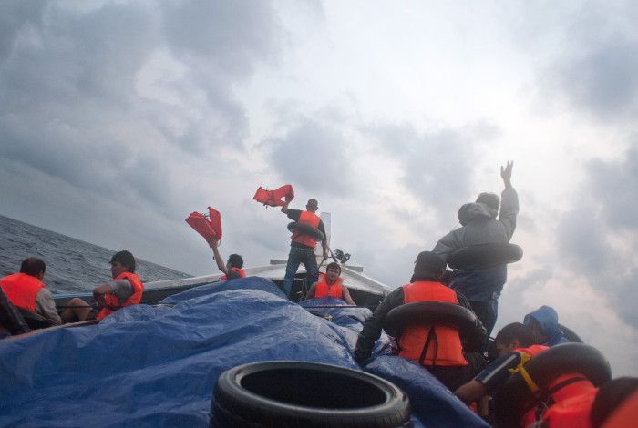 Figures on a boat, in life-vests, holding life vests aloft, against an overcast sky