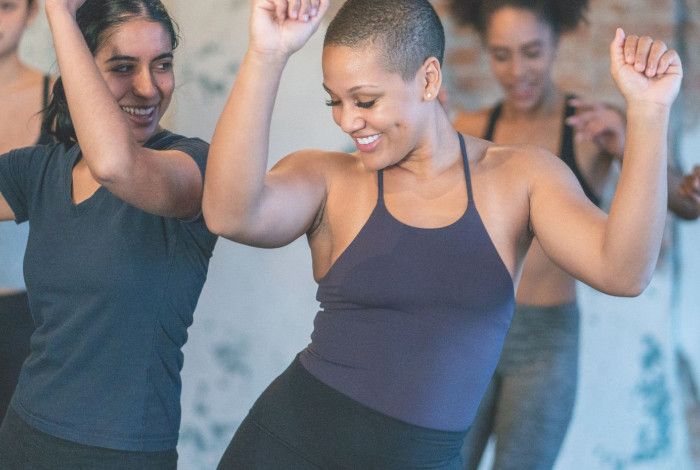Women dancing joyfully in a studio, smiling and raising their arms during a group fitness session.