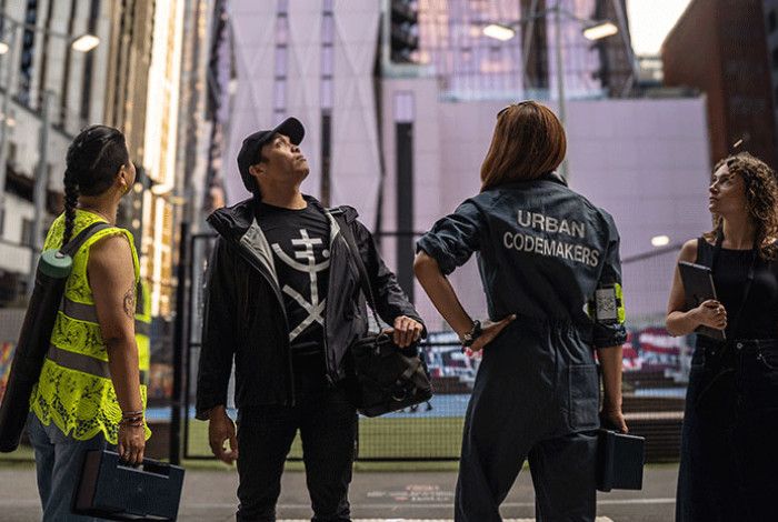 4 people standing in an urban environment. One wears a jumpsuit that says  Urban code makers. 