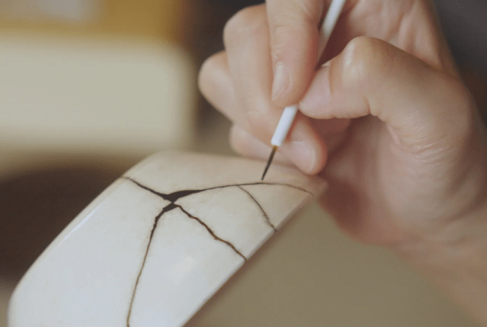 Hand repairing pottery with kintsugi, highlighting cracks with dark lacquer.