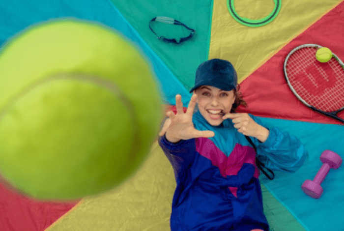 A person in sportswear reaches for a large tennis ball on a colorful mat surrounded by sports equipment