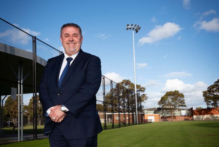Man in a dark suit stands on a sports ground under a large light tower