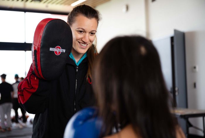 A smiling woman holds a boxing pad in front of someone standing with their back to the camera