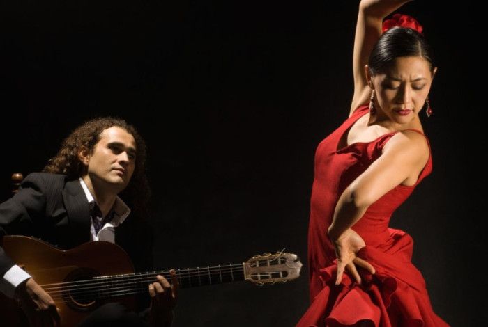 Male playing guitar while a lady in a red dress dances in front of him.