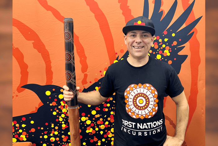 Person holding a decorated didgeridoo against a vibrant Indigenous-style backdrop.