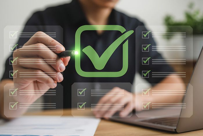 Hand marking a green tick on a digital checklist beside a laptop.