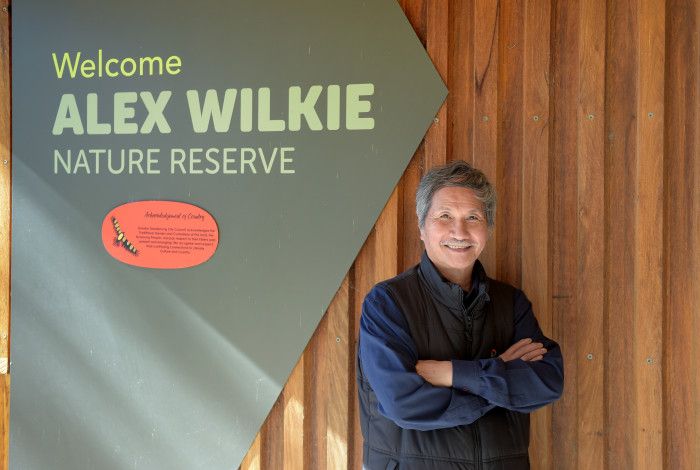 A smiling man with his arms crossed next to a sign that reads Welcome Alex Wilkie Nature Reserve