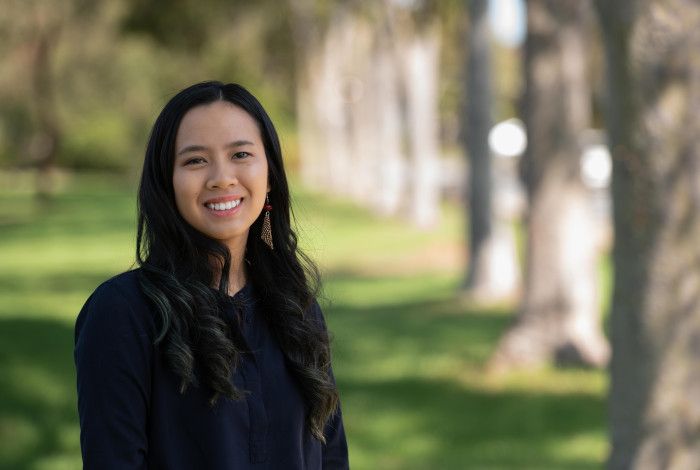 A woman with long dark hair smiles at the camera while standing in a park location