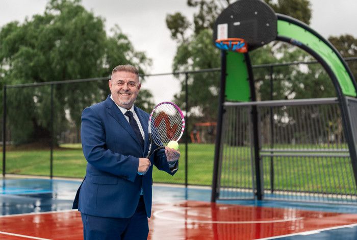 Smiling man posing on a sports court while holding a tennis racquet and tennis call.