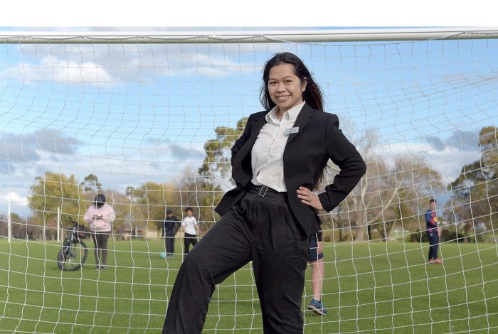 A smiling woman dressed in a black suit and white shirt stands in front of a soccer goal with one hand on her hip