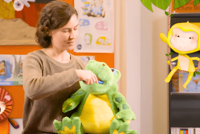 A woman holding a toy dinosaur and brushing his teeth against the background of bookshelves and toys.