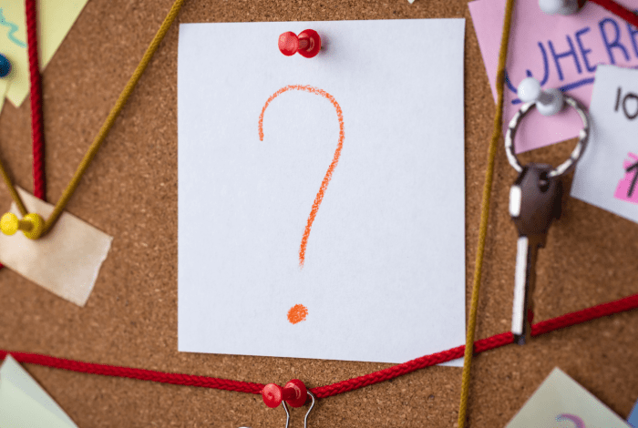 A question mark on a piece of paper pinned to a cork board with strings, keys and papers pinned around it. 