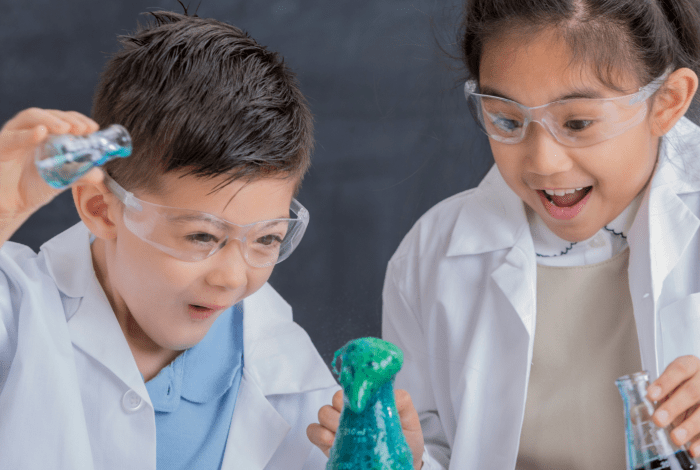 A boy and a girl dressed in white lab coats, wearing safety goggles, holding glass flasks and performing chemical experiments.
