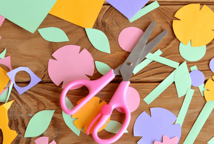 Pink scissors surrounded by colourful paper flowers, leaves and craft cut-outs on a wooden table.