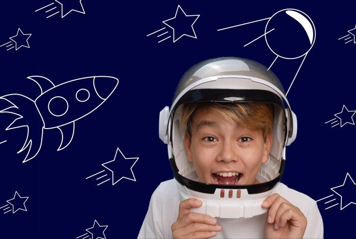 A boy dressed as an astronaut, laughing with excitement, standing in front of a drawing depicting space, rockets and stars.