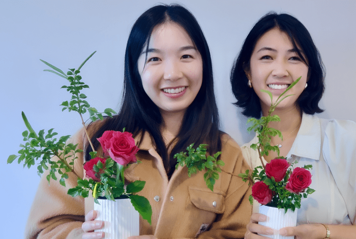 Two Japanese women standing together, facing the camera and holding their ikebana flower arrangements. 