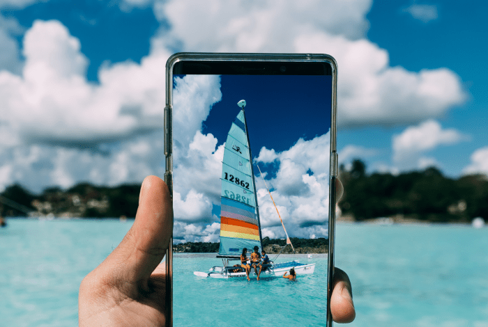 A smartphone frames a colorful sailboat on clear turquoise water beneath a bright sky filled with large white clouds.