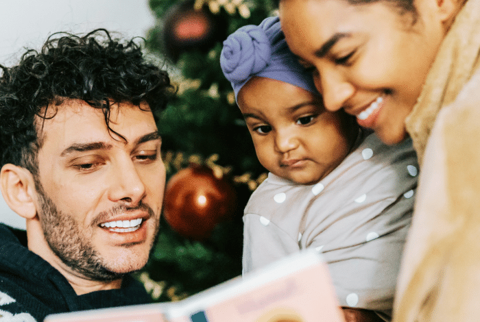 A multicultural family of three standing in front of a Christmas tree and reading a book.
