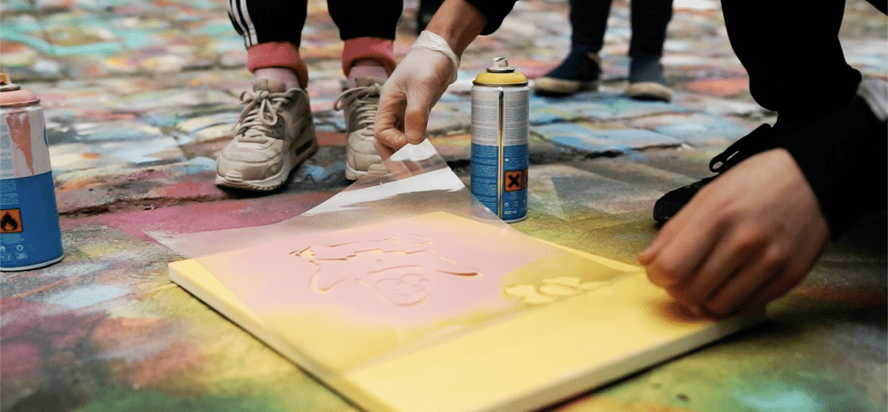 Hands placing a stencil down onto a canvas with onlookers in the background.
