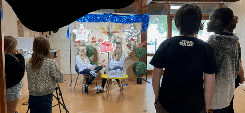 A group of kids ages between 10 - 17 inside a studio. Some kids are operating the camera and some kids are being filmed conducting an interview.