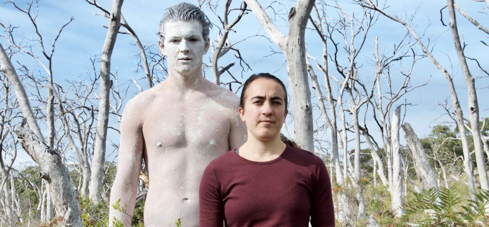 Two performers, one covered in white body paint, in front of bare trees