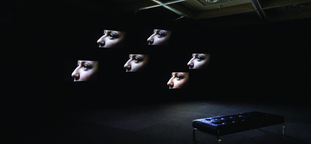 Installation view of Vernon Ah Kee's work, Lullaby, featuring 6 profiles of faces displayed on screens in a darkened room