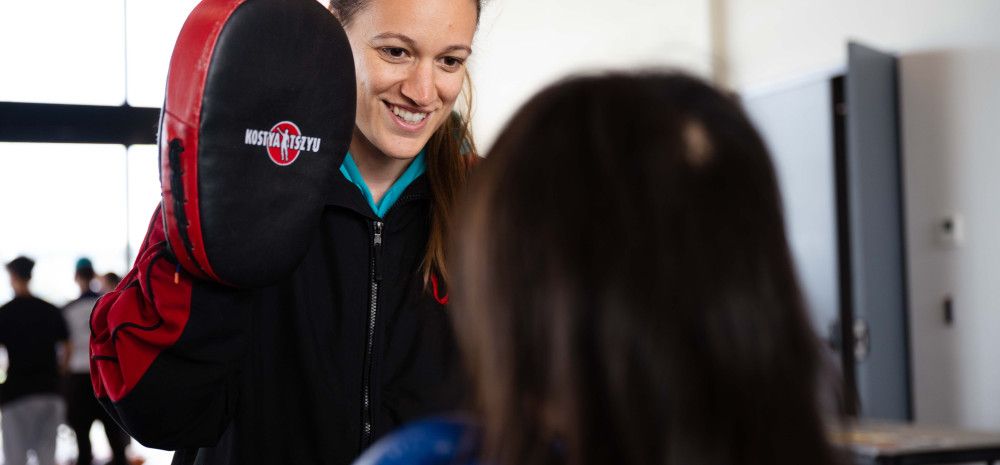 A smiling woman holds a boxing pad in front of someone standing with their back to the camera