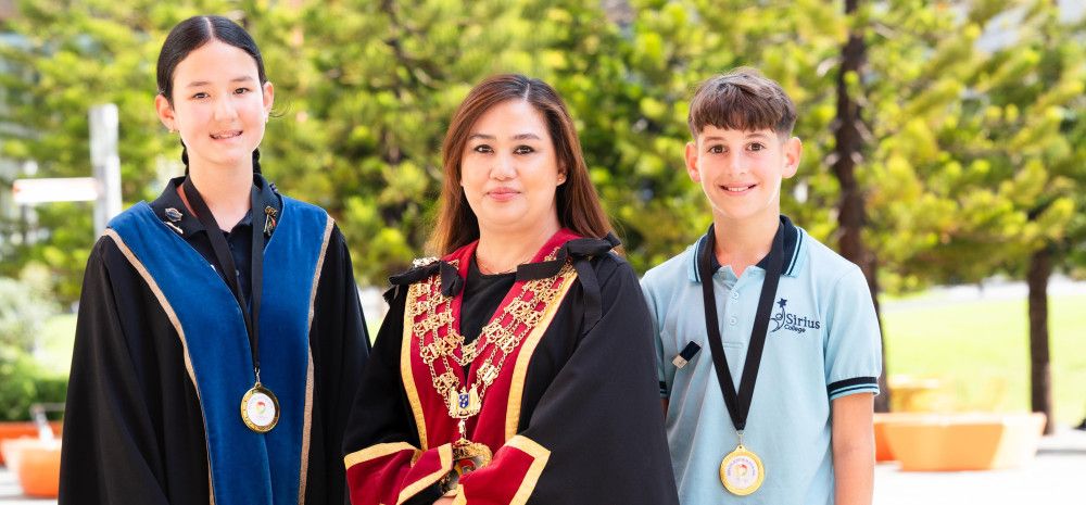 Two smiling children stand either side of a woman dressed in red and black robes
