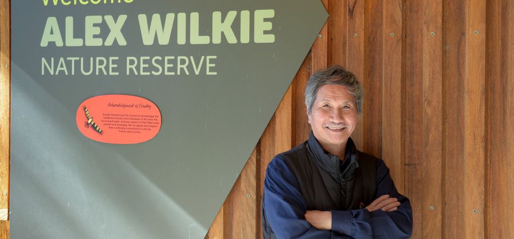 A smiling man with his arms crossed next to a sign that reads Welcome Alex Wilkie Nature Reserve