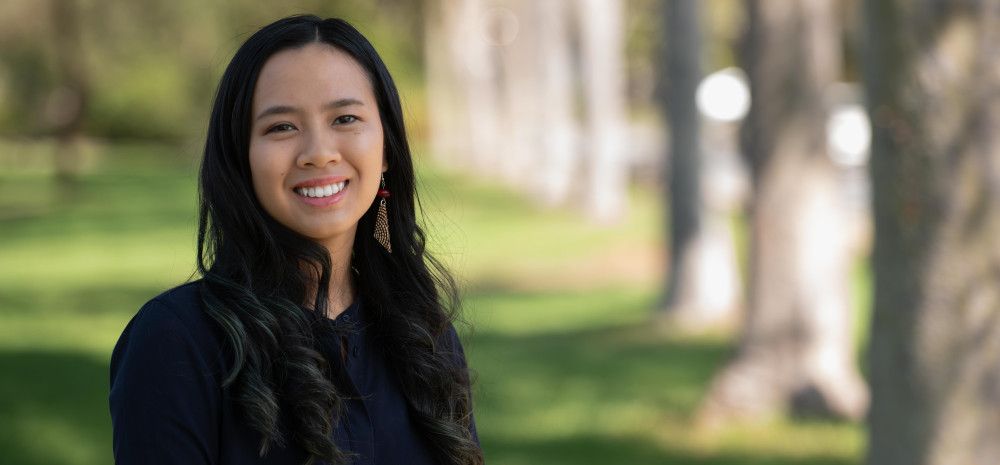 A woman with long dark hair smiles at the camera while standing in a park location