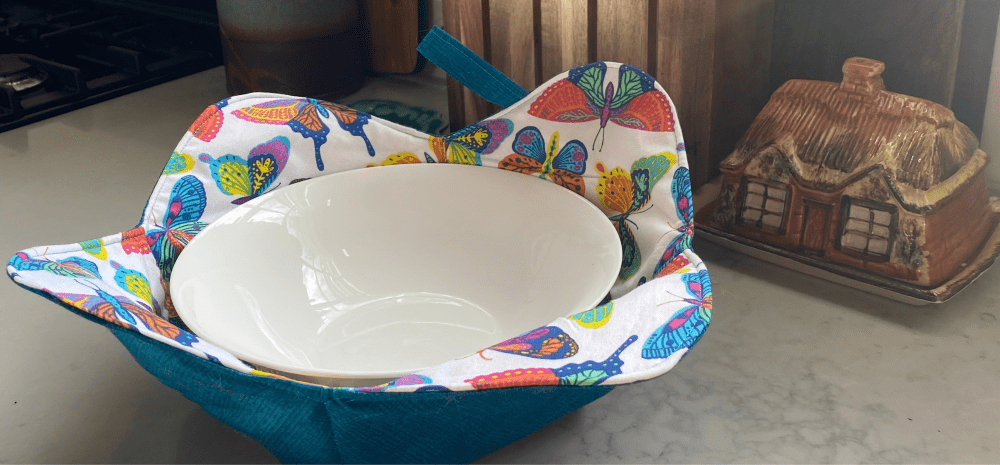 A white bowl in a colourful fabric bowl cosy next to a little china house model. 