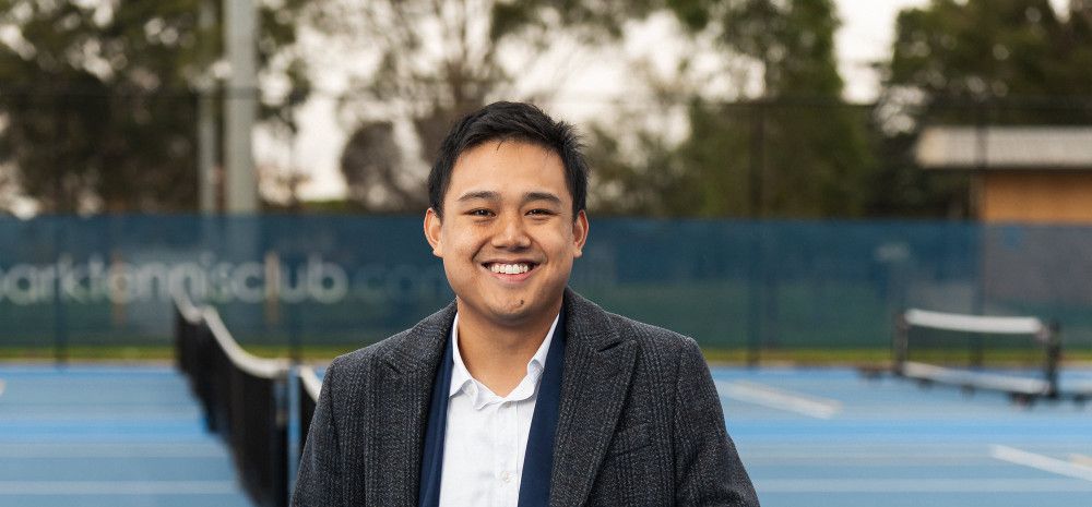 A smiling man leans on a tennis net.
