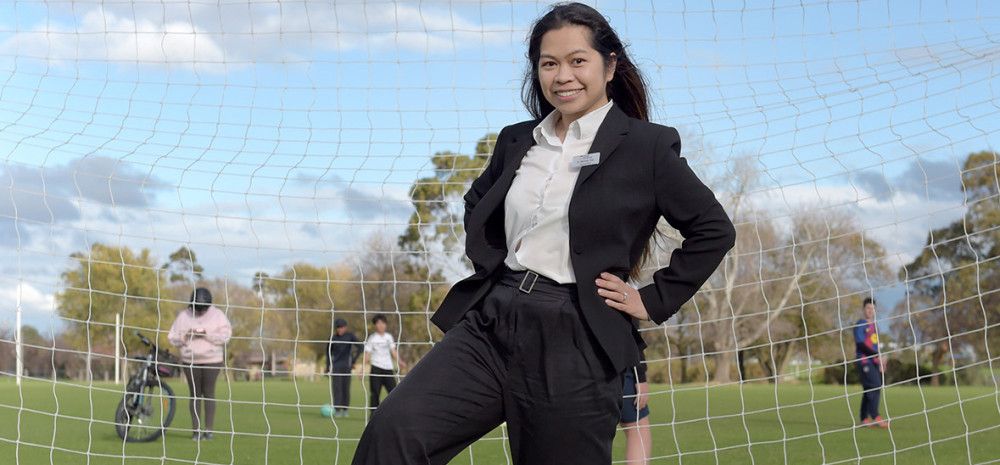A smiling woman dressed in a black suit and white shirt stands in front of a soccer goal with one hand on her hip